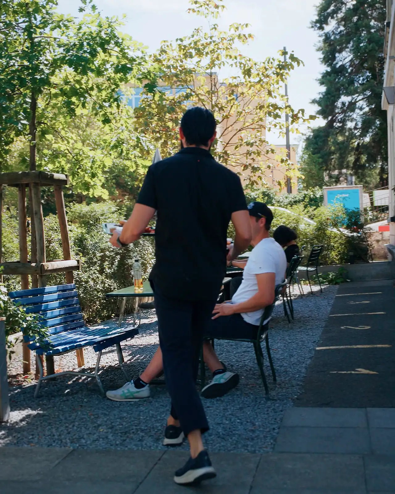 Unsere Gartenterrasse im Sommer: Tische auf Kies unter Bäumen, links eine blaue Bank, ein Gast am Tisch, ein Mitarbeiter bringt ihm die Bestellung
